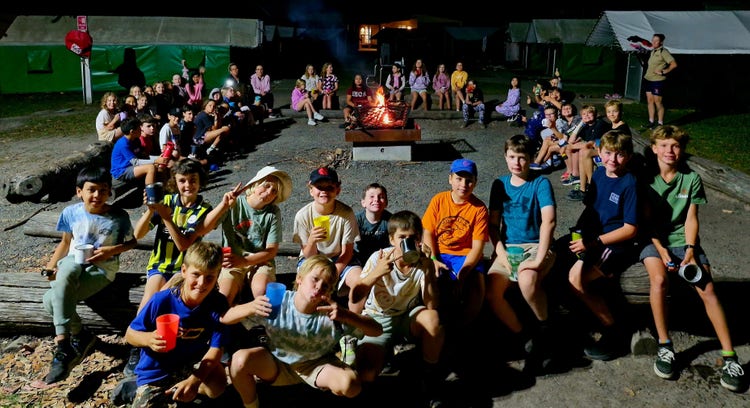 Group fo year 4 students at a year 4 camp. The students rae sitting around a camp fire with cups of hot chocolate.